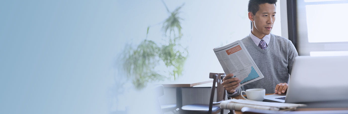 Asian guy using laptop while holding economic newspaper